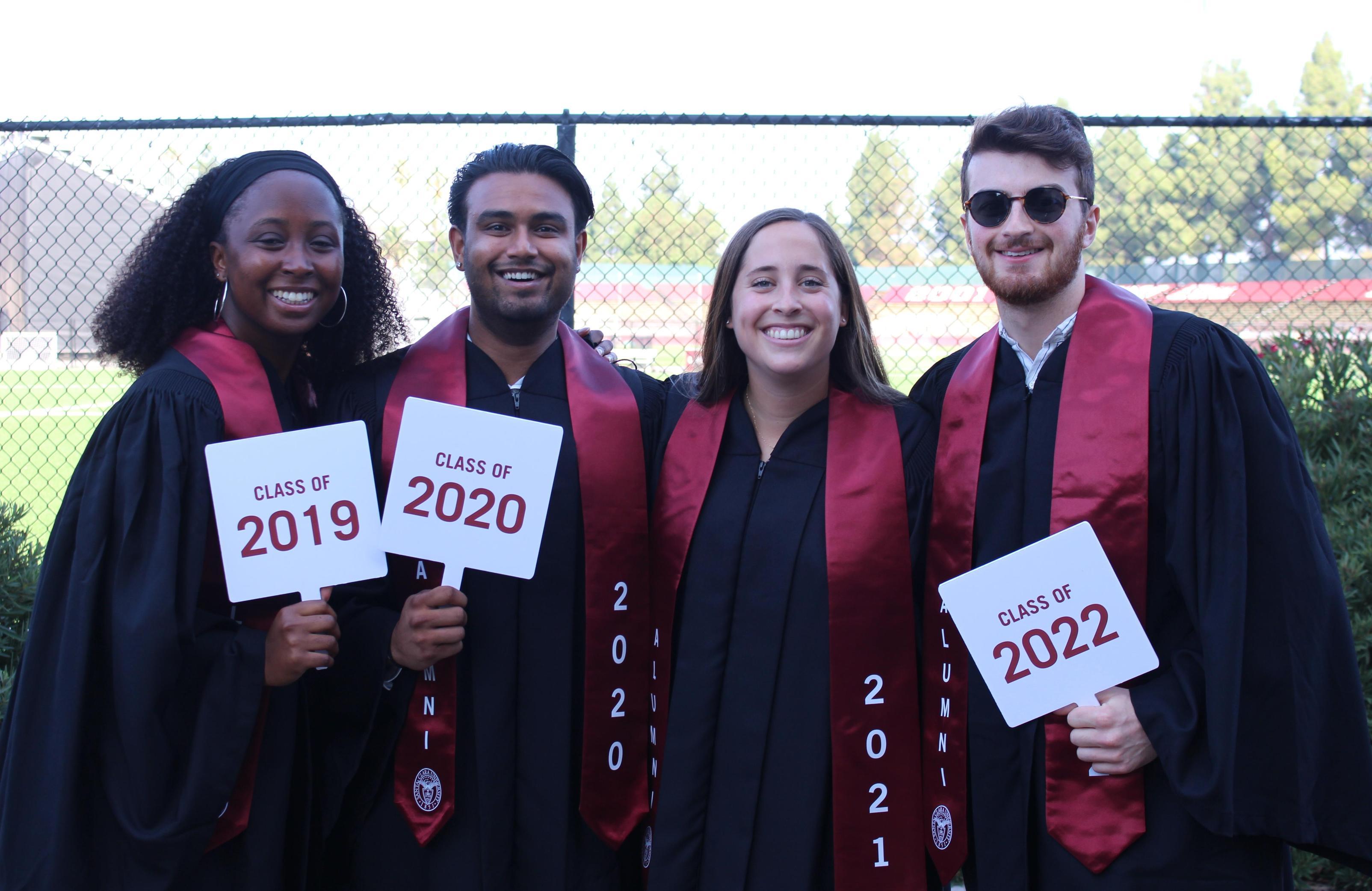 Four graduates holding Hire Me! signs and wearing caps and gowns.