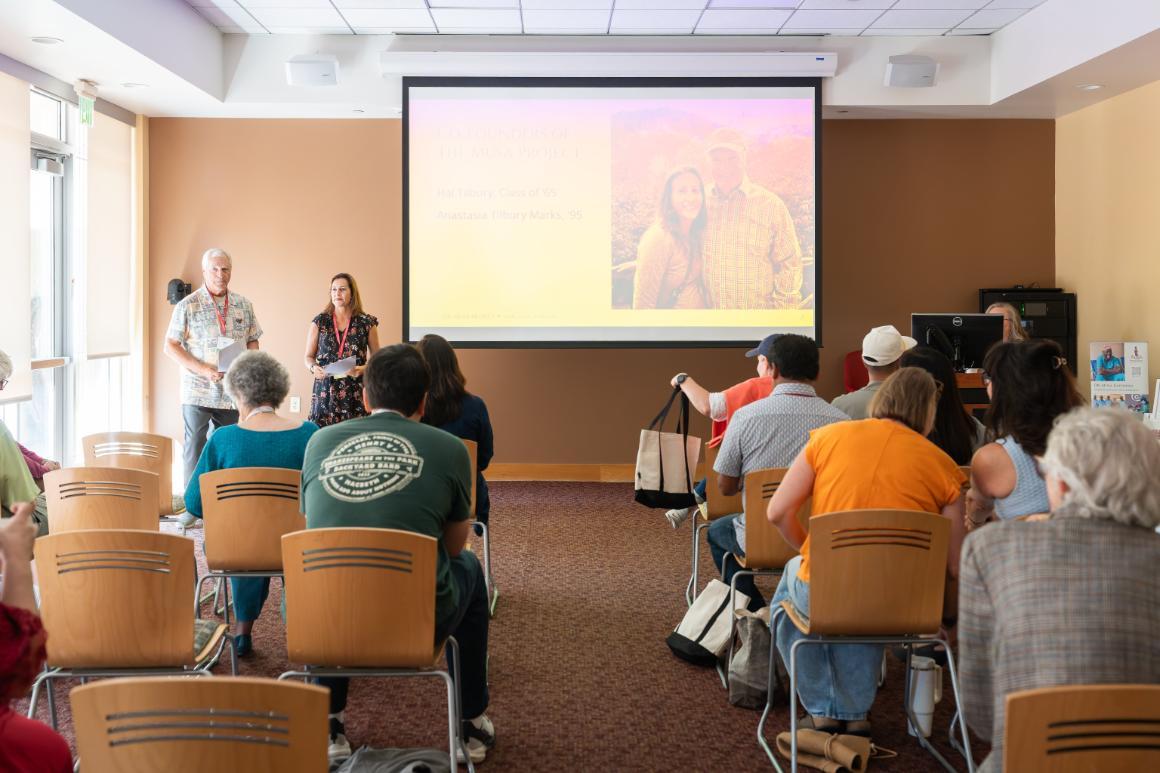 A man and a woman stand in front of a seated audience, and next to a projector screen with their photo on it, along with text reading Co-founders of The Musa Project. Hal Tilbury, Class of '65. Anastasia Tilbury Marks '95.