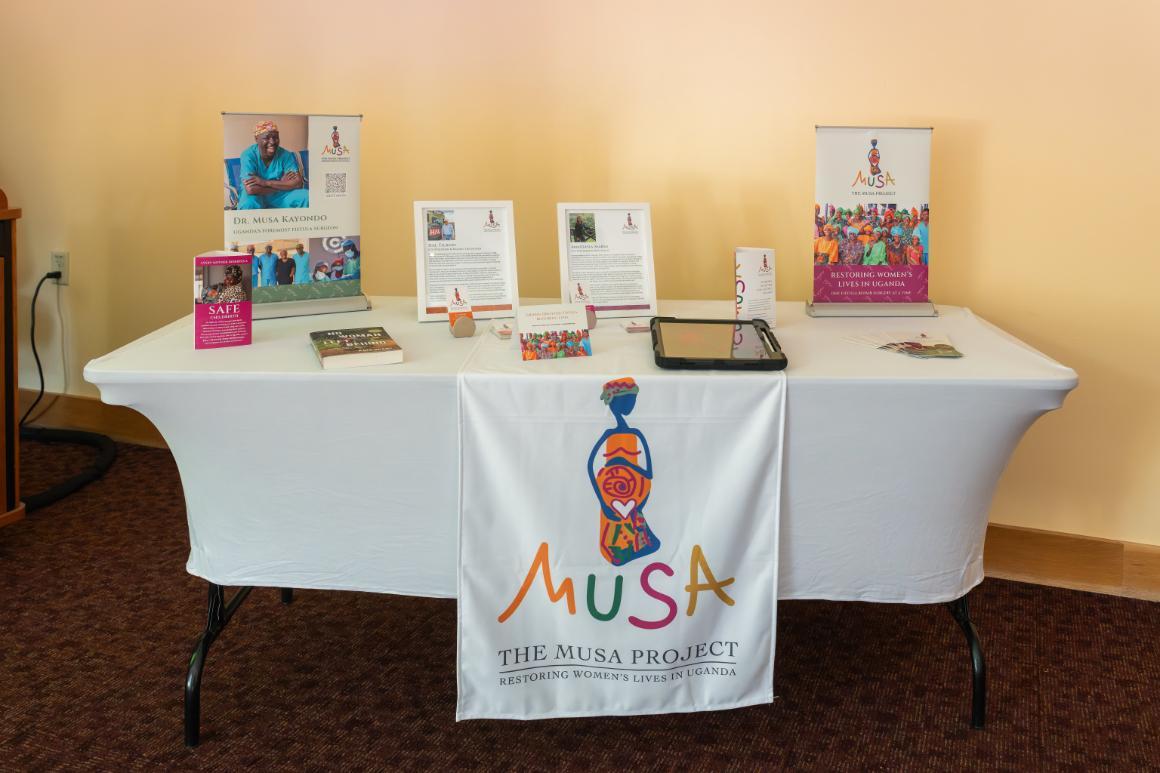 A table with a white tablecloth and a table runner reading Musa. The Musa Project. Restoring Women's Lives in Uganda. The tabletop holds several stacks of fliers and some small, pop-up banners with more information about The Musa Project.