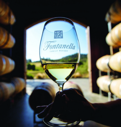 Wine glass with Fontanella logo near winery barrels and vineyard view.