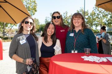 Four people posing together at an outdoor event near a red table.