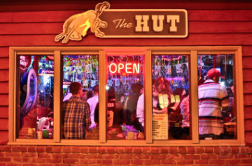 A group of people as seen through a set of five windows. A sign above the windows reads The Hut with a bucking bronco next to the text. A smaller, neon sign in the center window reads Open.