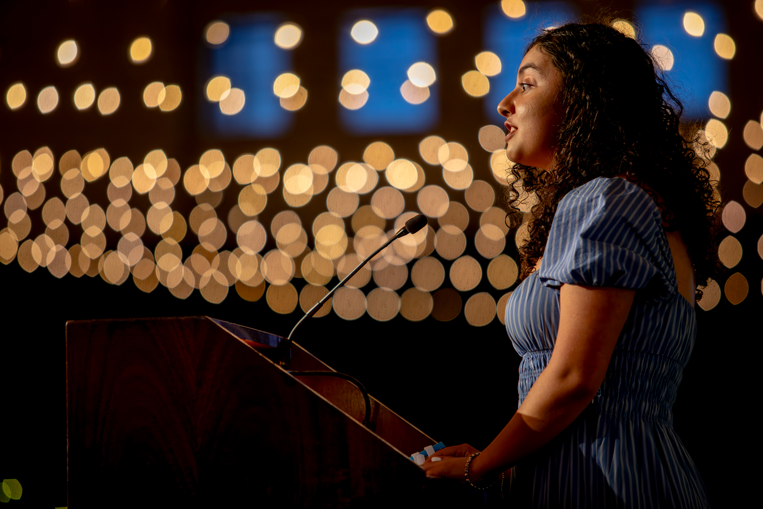 A young woman speaking at a podium with bistro lights in the background