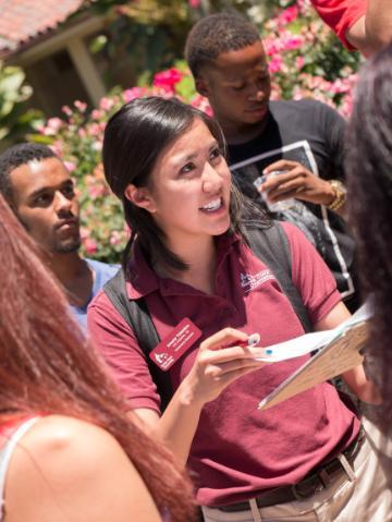 Group of people outside, one person speaking and holding a clipboard.