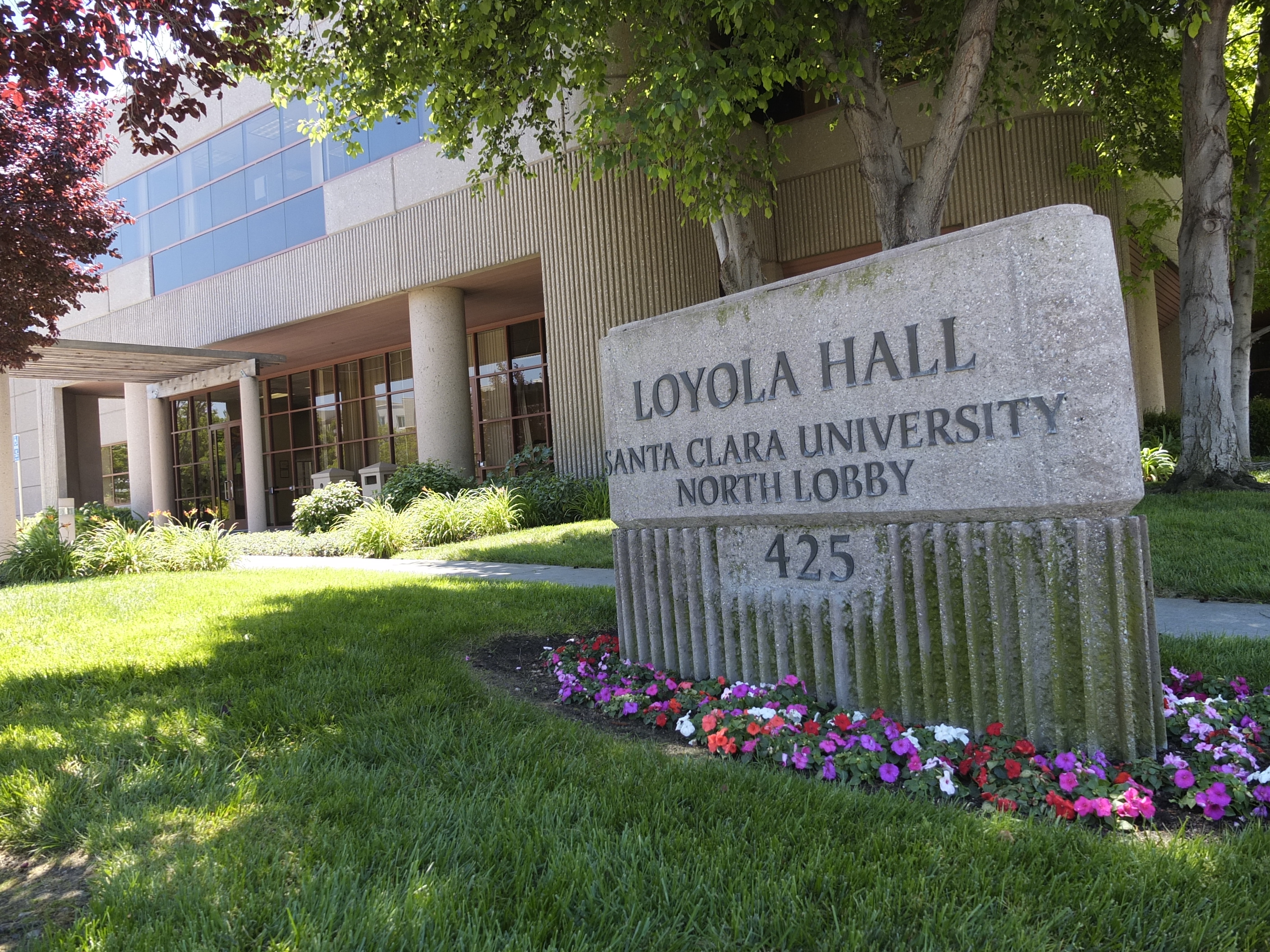 Loyola Hall North Lobby building sign with grass, flowers, and trees in the background.