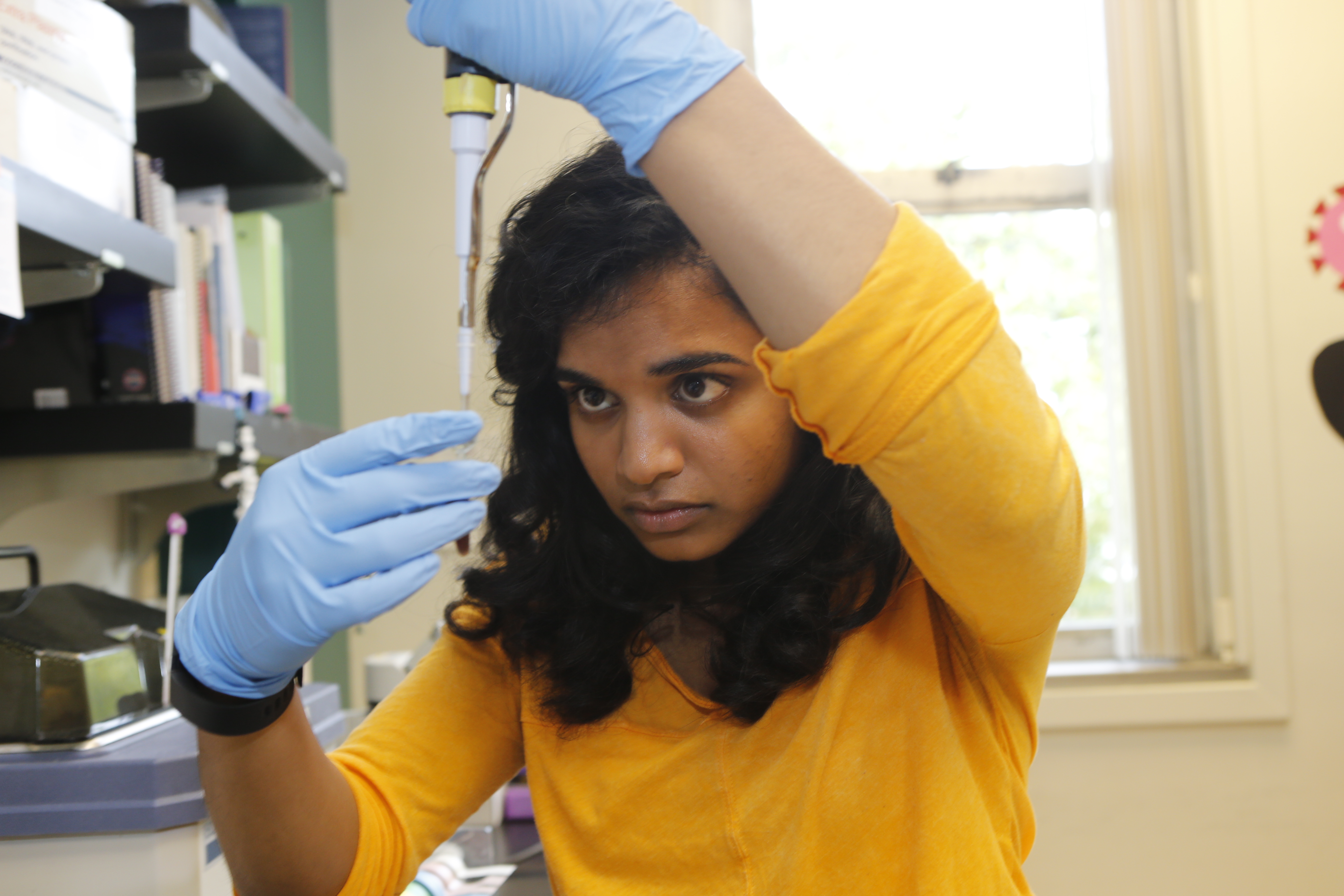 Student working in a lab, focused on a pipette and vial held at eye level.