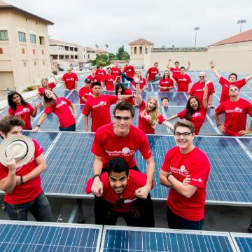 Students wearing matching red shirts gathered around solar panels smiling. Links to Civil, Environmental, and Sustainable Engineering MS page.