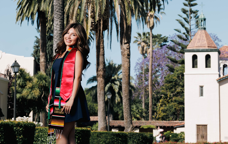SCU Graduate Raechel Guevara stands in front of the mission church wearing graduation cords and garlands. image link to story