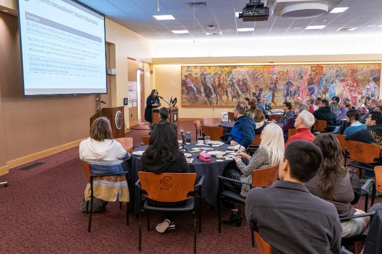 Sha Duncan Smith presents at the front of a large room as attendees listen from their tables. Photo by Jim C