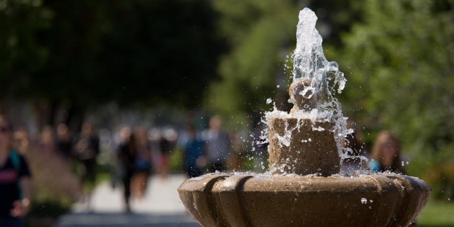 A bubbling fountain with people walking on campus in the background