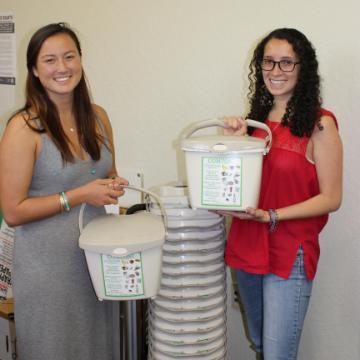 Two women holding compost bins in a room titled Villas Composting.