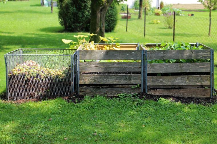 Three-bin compost system in a grassy yard with trees and a fence in the background.