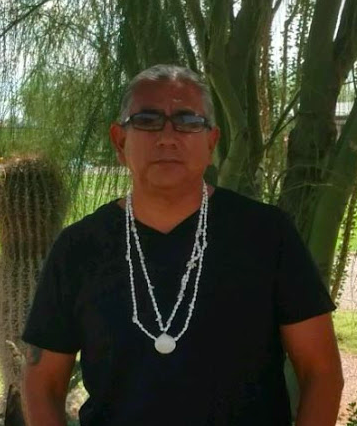 A person wearing glasses and necklaces stands in front of trees.