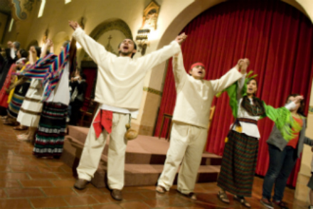 People in traditional attire performing on stage with red curtains in the background.