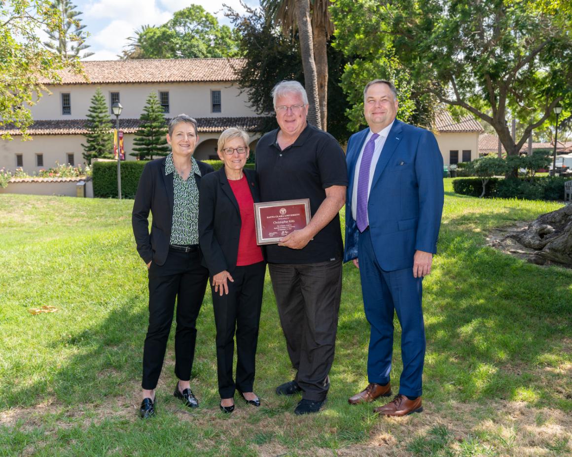 Four people standing outdoors, one holding an award plaque titled Kitts award. image link to story