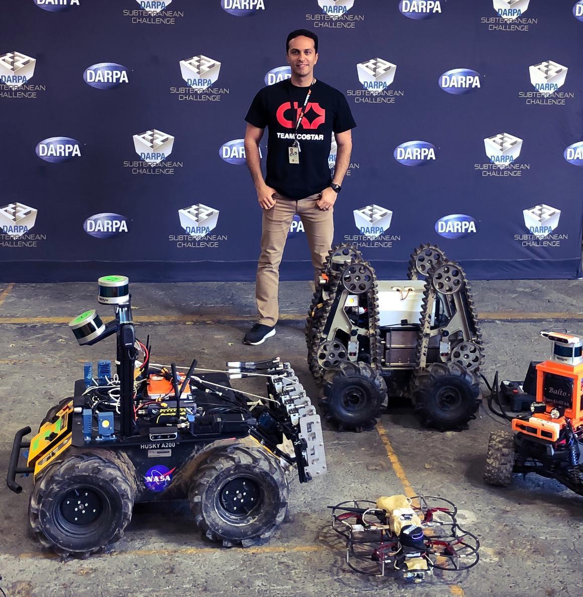 Kamak Ebadi standing in front of a DARPA sign with a variety of robots at his feet image link to story