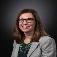 A person wearing glasses, a green blouse, and a light jacket, smiling against a dark background.