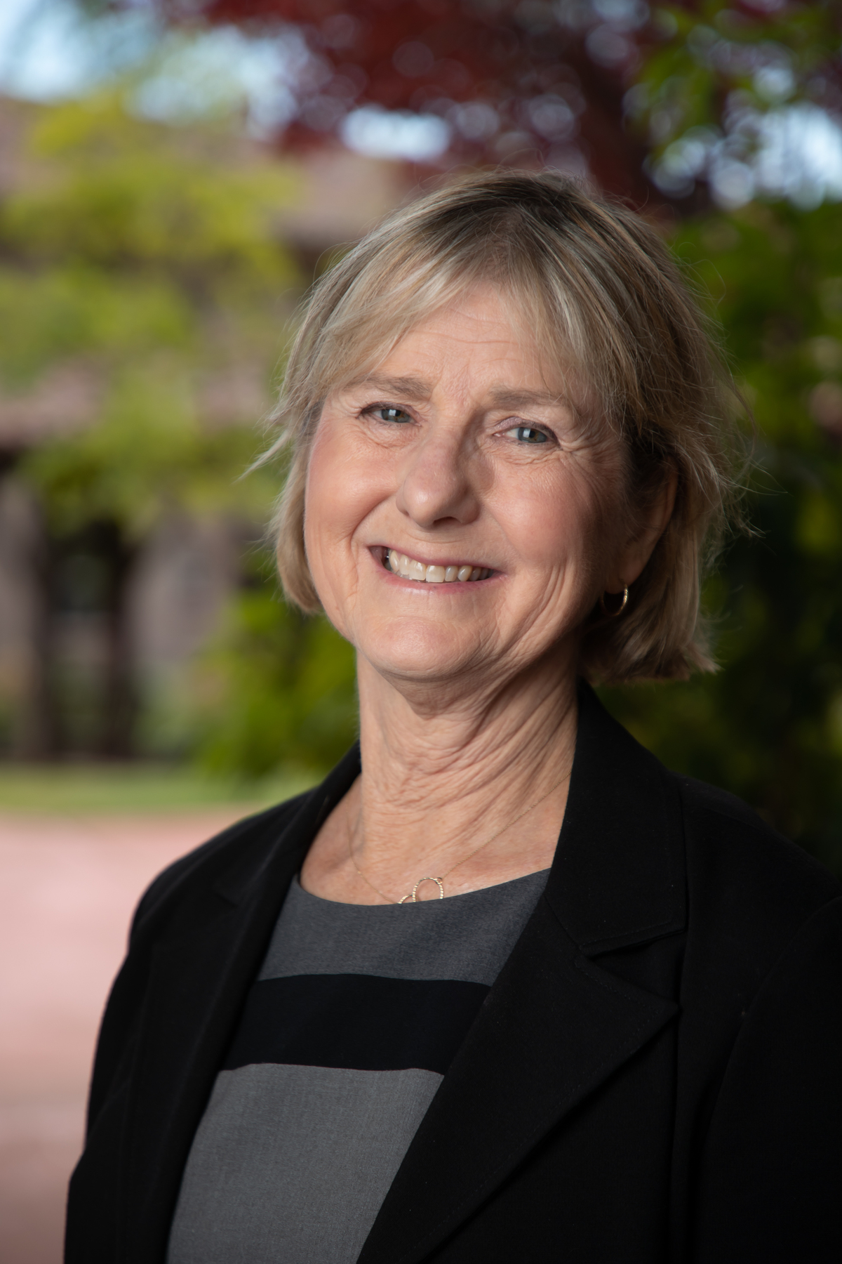 A person in professional attire smiling outdoors with trees and buildings in the background.