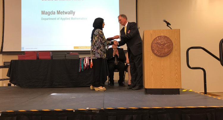 A person receiving an award on stage from another person, with Magda Metwally projected behind them.