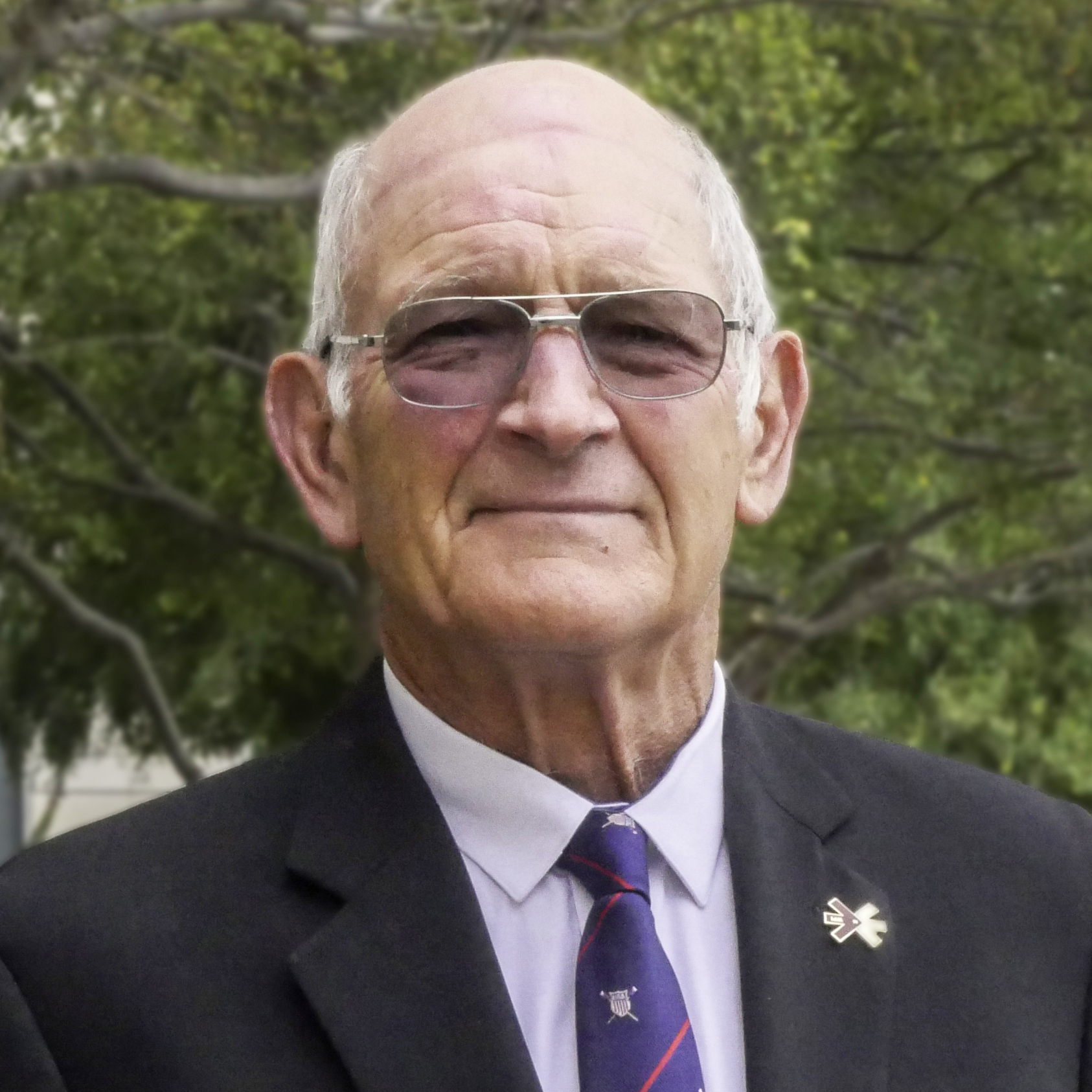 A man wearing glasses, a suit, and a tie, with trees in the background. image link to story