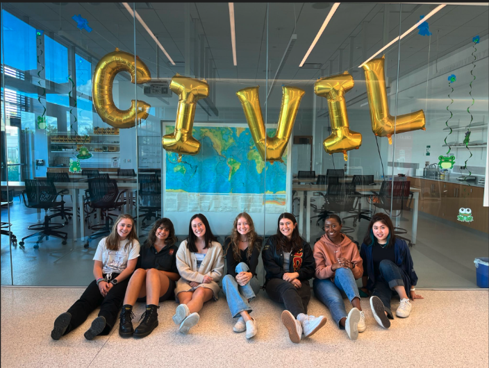 Seven female students sitting on the floor in front of the Geotech Lab with big balloons spelling out CIVIL taped to the glass.