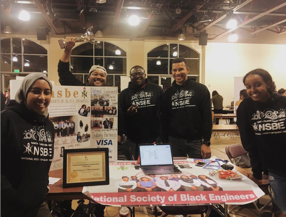 Group posing at NSBE Involvement Fair booth with displayed items and poster.