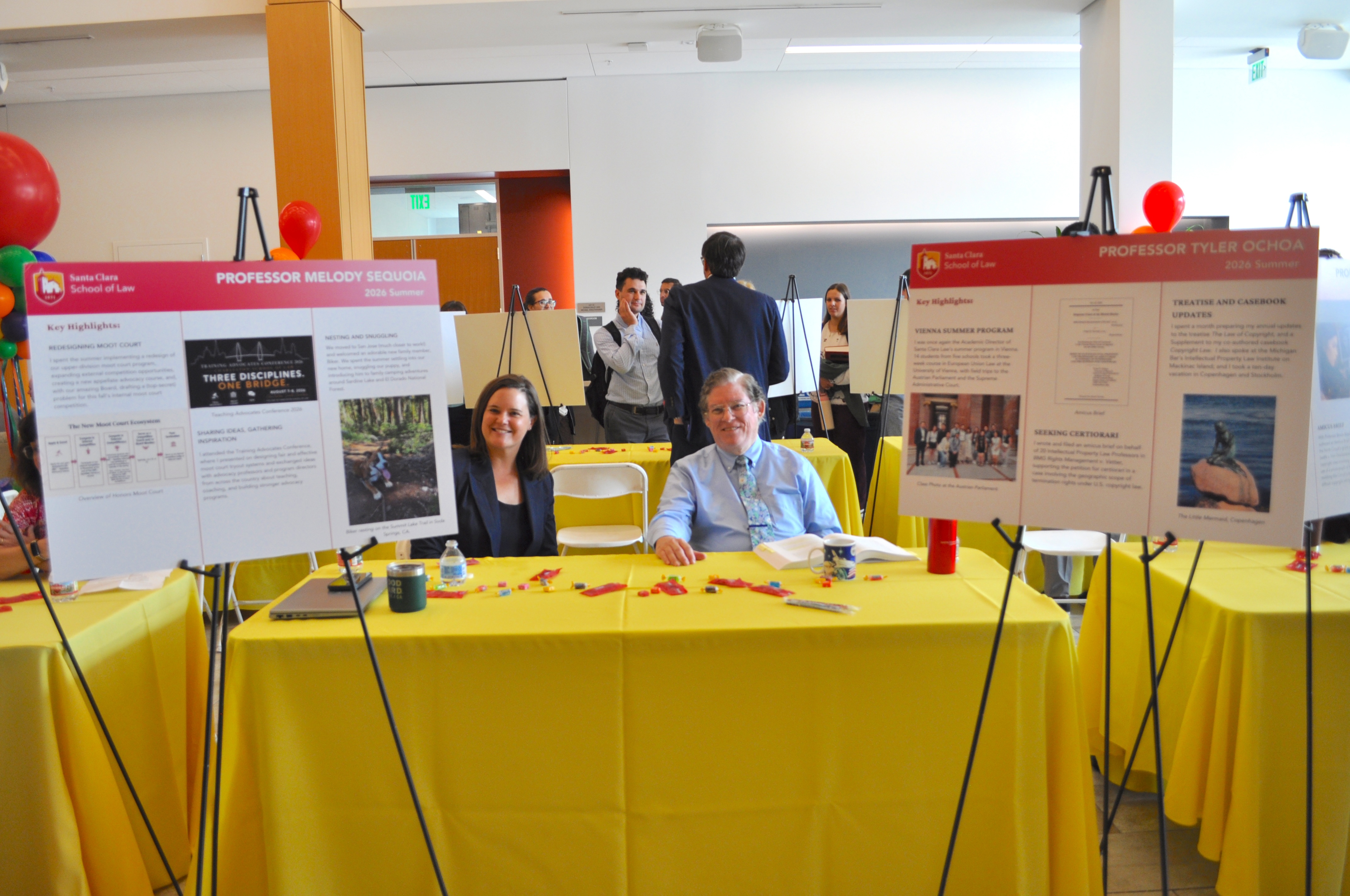 Two professors sit behind table with poster boards on either side smiling at camera